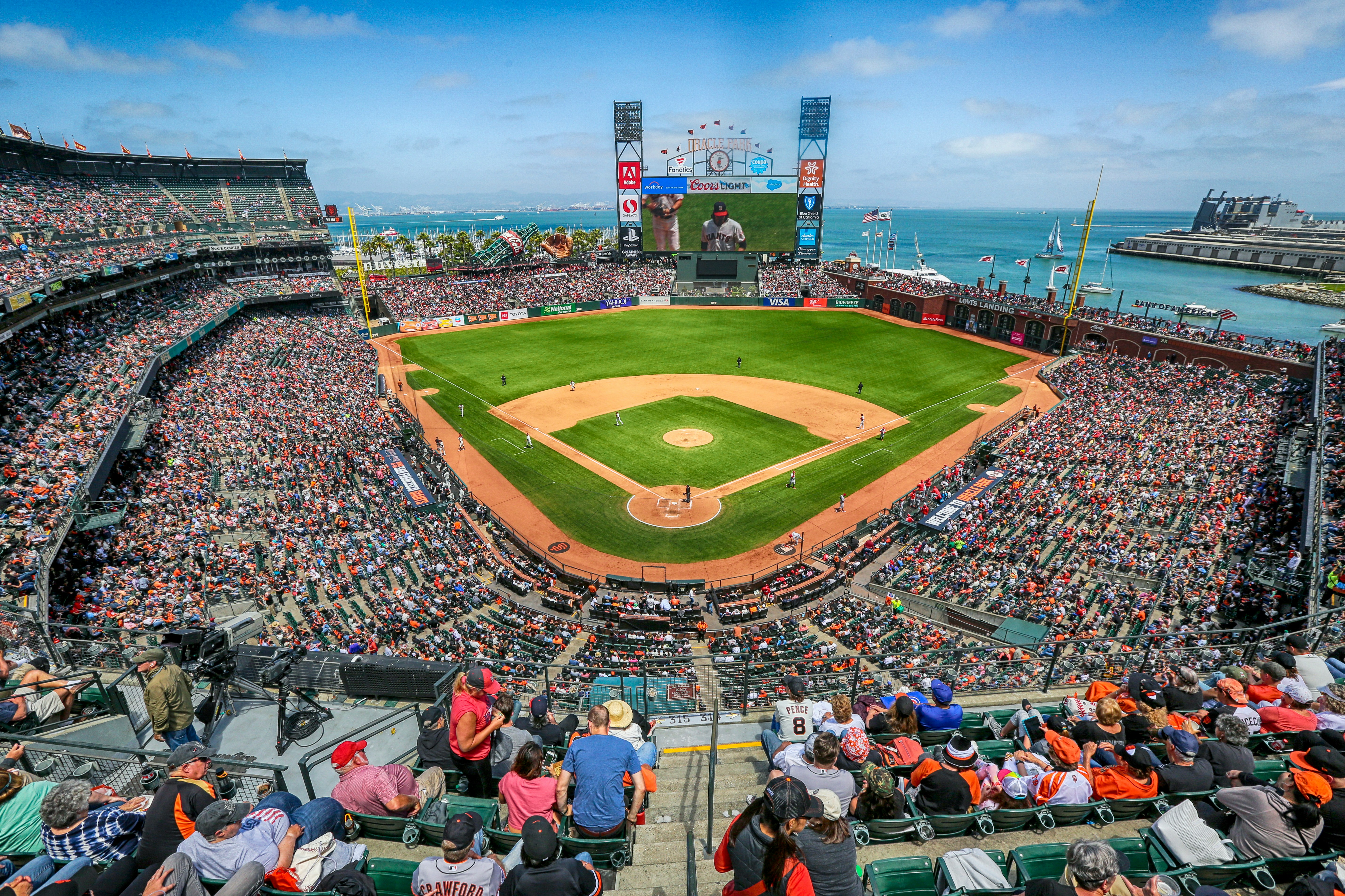 Packed baseball stadium with ocean view, green field, and large video scoreboard