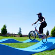 Mountain biker in black gear jumping bicycle on blue pump track with green landscape