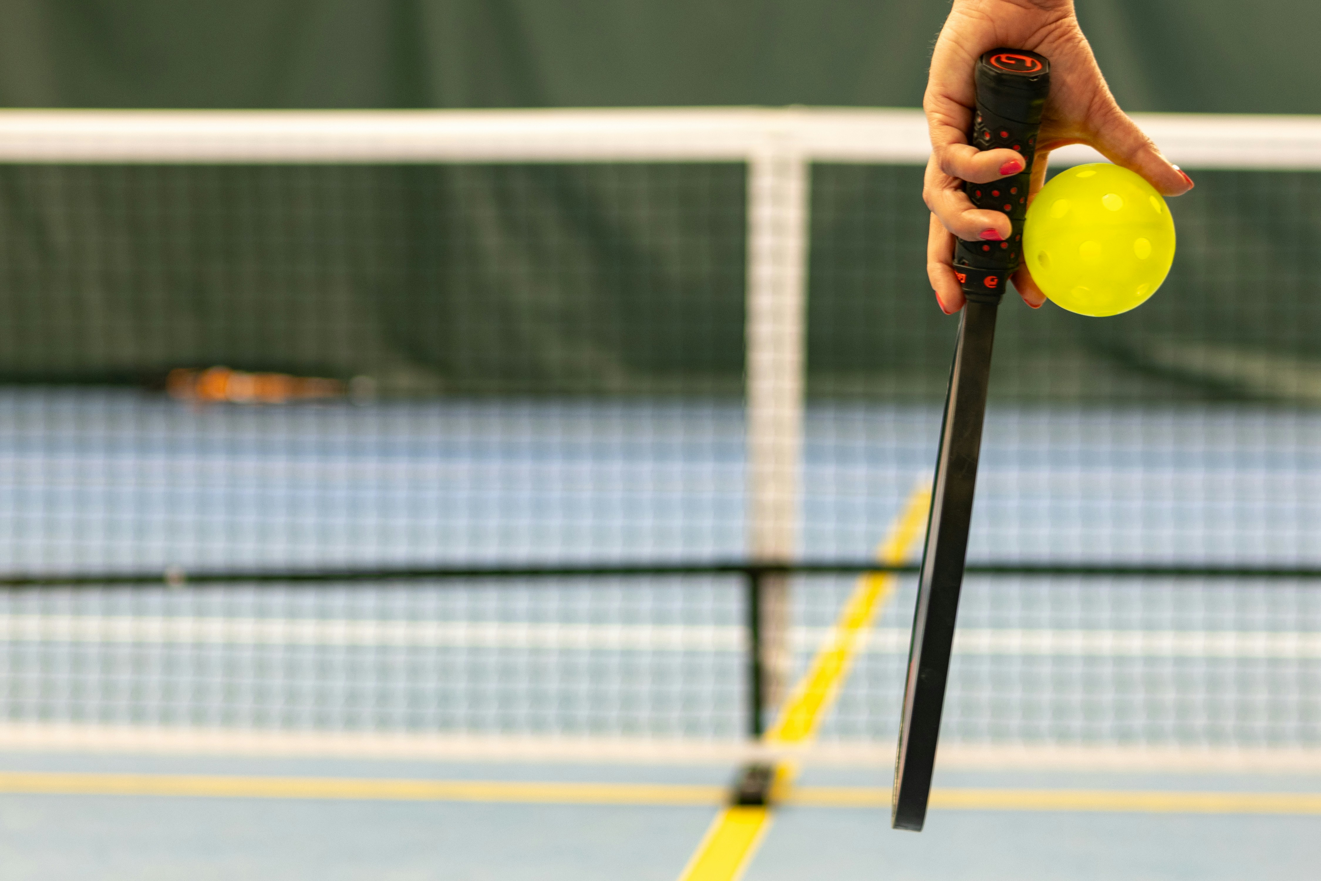 Hand holding yellow pickleball and paddle near court net