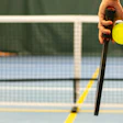Hand holding yellow pickleball and paddle near court net