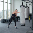 Woman punching heavy bag in modern gym with equipment and city view through windows