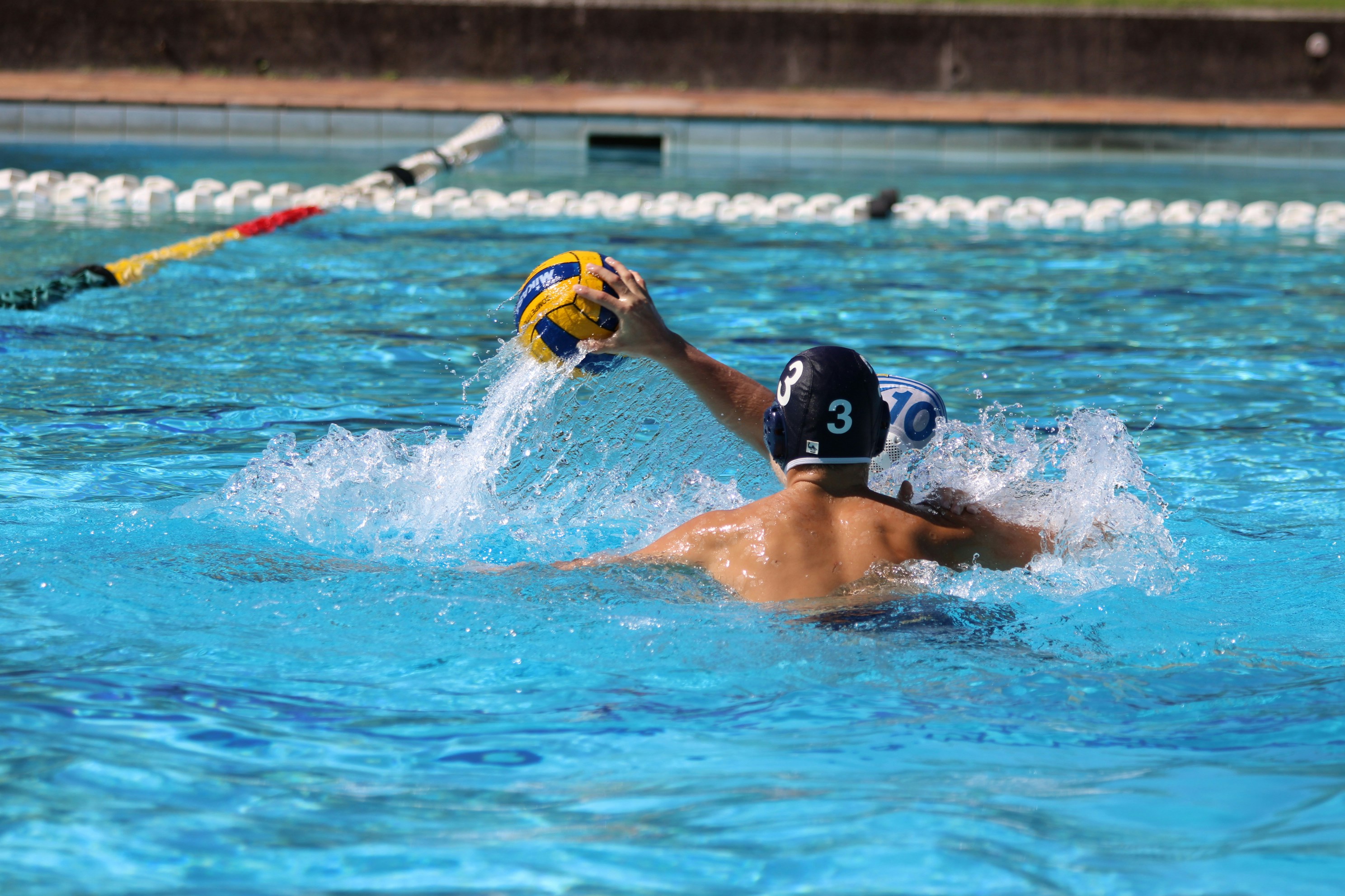 Water polo player in black cap with number 9 throws yellow ball during competitive match in pool