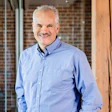 Professional headshot of a smiling middle-aged man wearing a light blue button-up shirt in a modern office setting