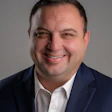 Professional headshot of a smiling man in navy blazer and white dress shirt with blue patterned pocket square