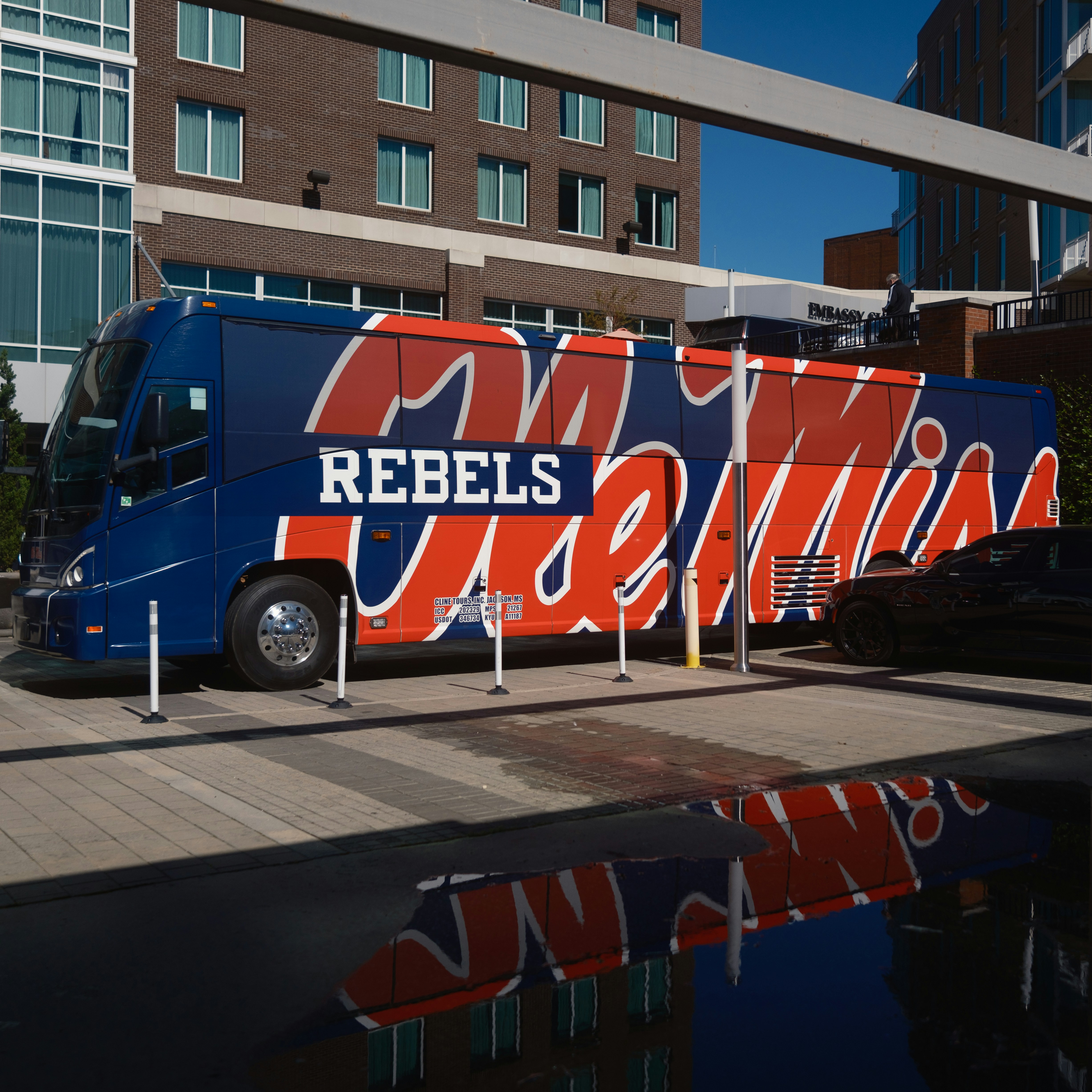 Blue and red branded tour bus with 'Rebels' text parked in urban plaza with modern buildings