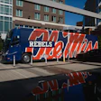 Blue and red branded tour bus with 'Rebels' text parked in urban plaza with modern buildings
