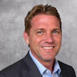 Professional headshot of a smiling man in dark blazer and light blue shirt against gray background