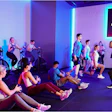 Group fitness class with participants on stationary bikes and running on floor under purple and blue neon lighting with Peloton branding on screen.