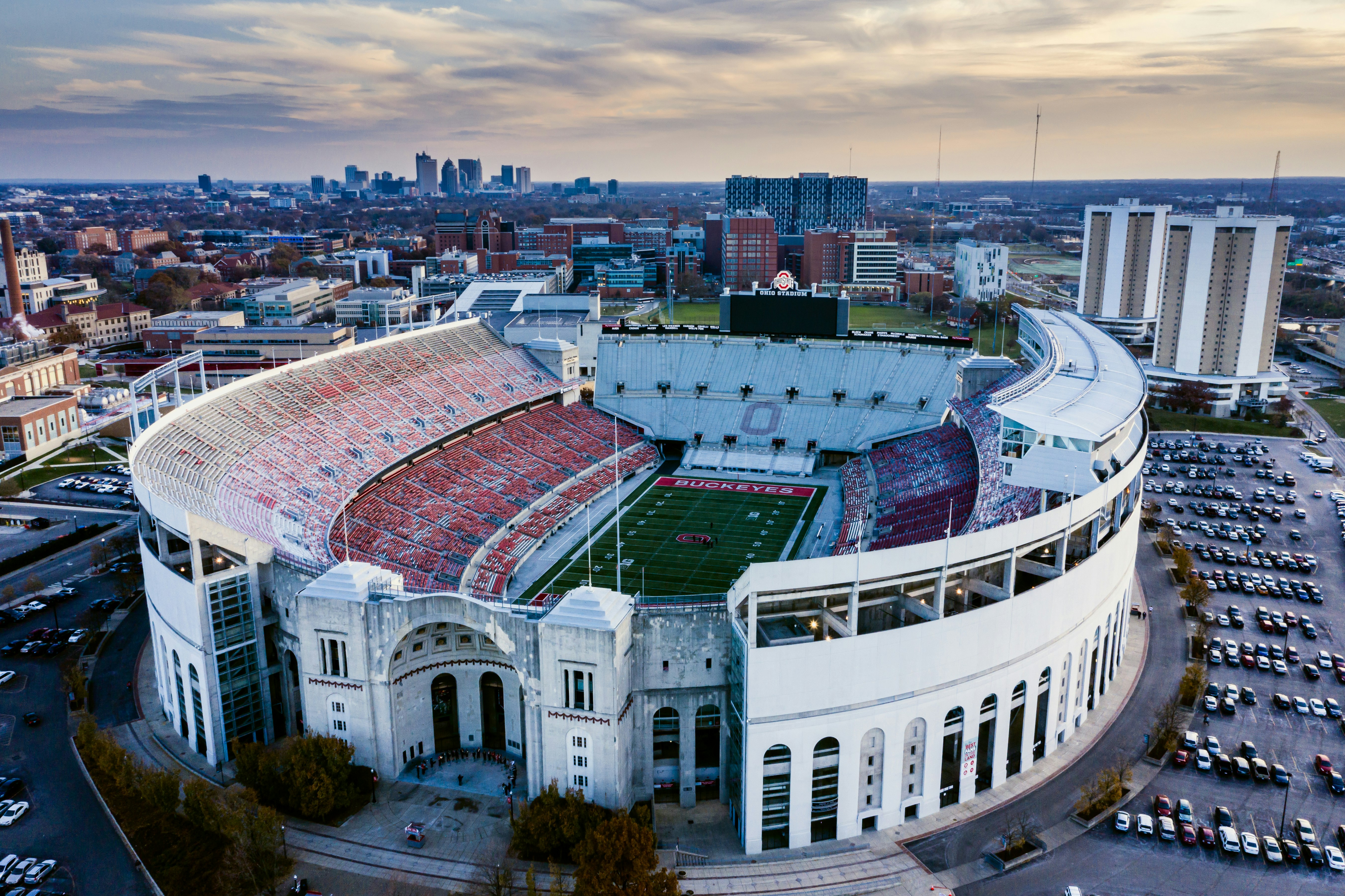 Aerial view of a large red and white sports stadium with curved architecture surrounded by urban cityscape and parking lots.