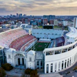 Aerial view of a large red and white sports stadium with curved architecture surrounded by urban cityscape and parking lots.