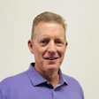 Professional headshot of a smiling man wearing a purple polo shirt against a neutral background