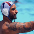 Male water polo player wearing red, white, and blue numbered cap in pool with arm extended