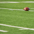 Red and brown football resting on green grass field with white yard line markings