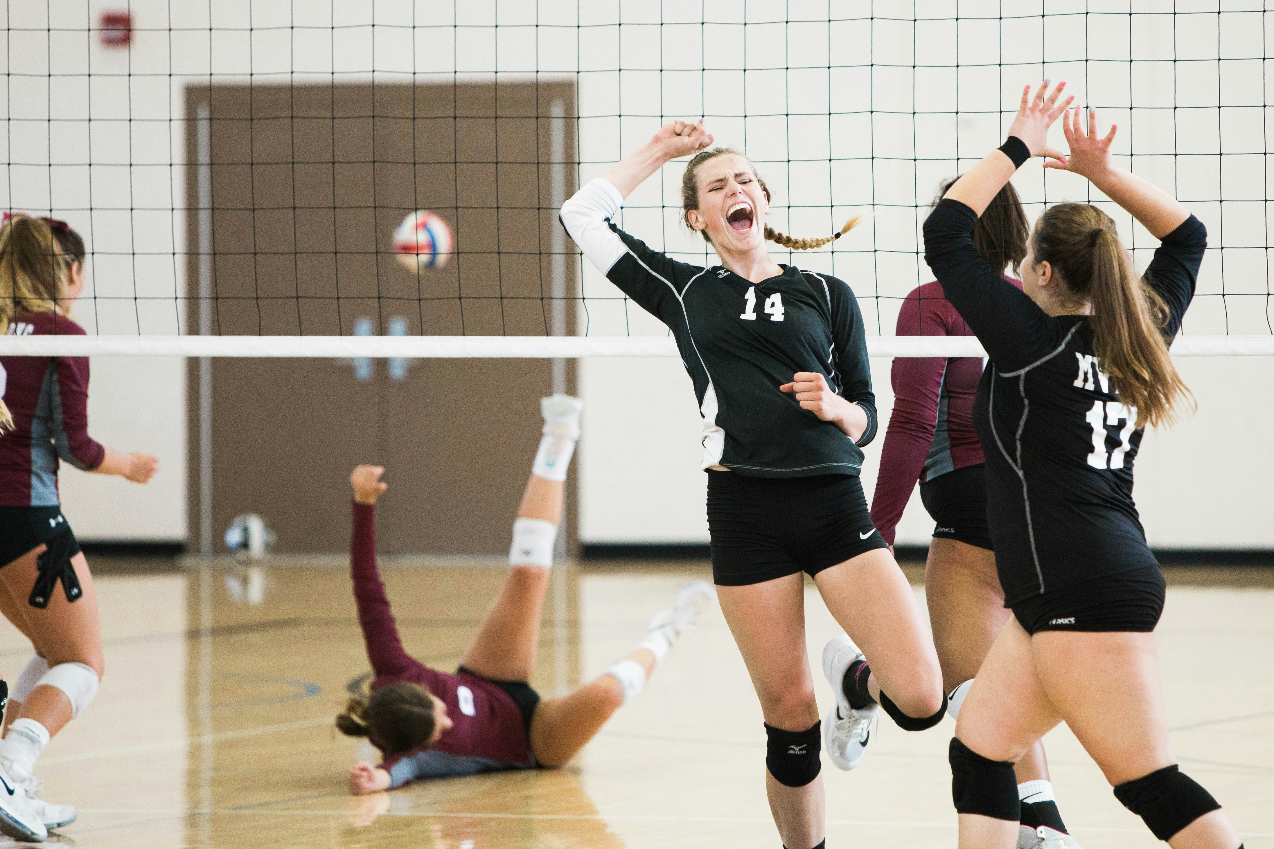 Young female volleyball players in black jerseys celebrate scoring a point during an indoor match at a gymnasium.