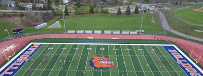 Aerial view of a high school football field with orange mascot logo, blue end zones, and surrounding athletic facilities and residential neighborhood