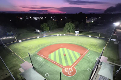 Illuminated baseball diamond at twilight with striped infield, surrounded by trees and town lights under purple sunset sky