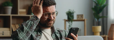 Bearded man wearing glasses and plaid shirt looking at smartphone in modern home office