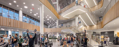 Modern multi-level fitness facility with exercise equipment, wooden railings, and people working out across interconnected floors.