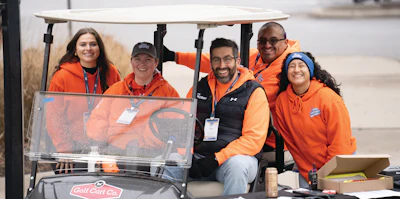 Five team members in orange apparel smiling together in a golf cart at an outdoor event
