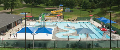 Aerial view of community water park with multiple pools, water slides, and blue shade structures surrounded by green lawn and trees