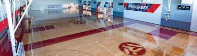 Modern gymnasium interior with polished wooden basketball court, maroon athletic logo, and progress signage on walls