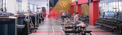 Modern gym interior with cardio equipment, weight training stations, and red accent walls with workout areas.