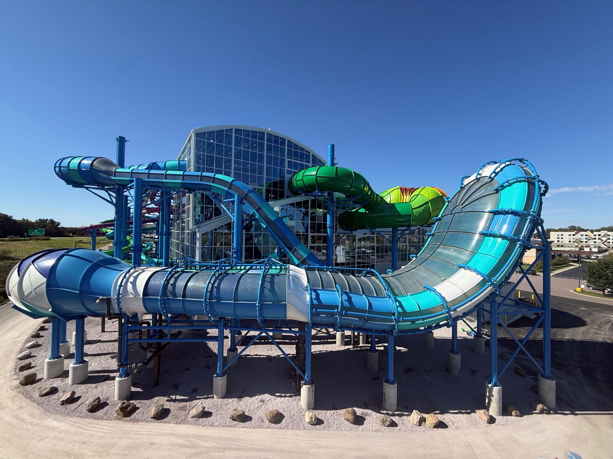 Colorful water park with multiple interconnected slides in blue, green, and cyan colors surrounding a central building structure under clear blue sky.