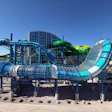 Colorful water park with multiple interconnected slides in blue, green, and cyan colors surrounding a central building structure under clear blue sky.