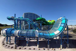 Colorful water park with multiple interconnected slides in blue, green, and cyan colors surrounding a central building structure under clear blue sky.