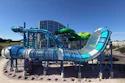 Colorful water park with multiple interconnected slides in blue, green, and cyan colors surrounding a central building structure under clear blue sky.