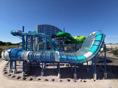 Colorful water park with multiple interconnected slides in blue, green, and cyan colors surrounding a central building structure under clear blue sky.