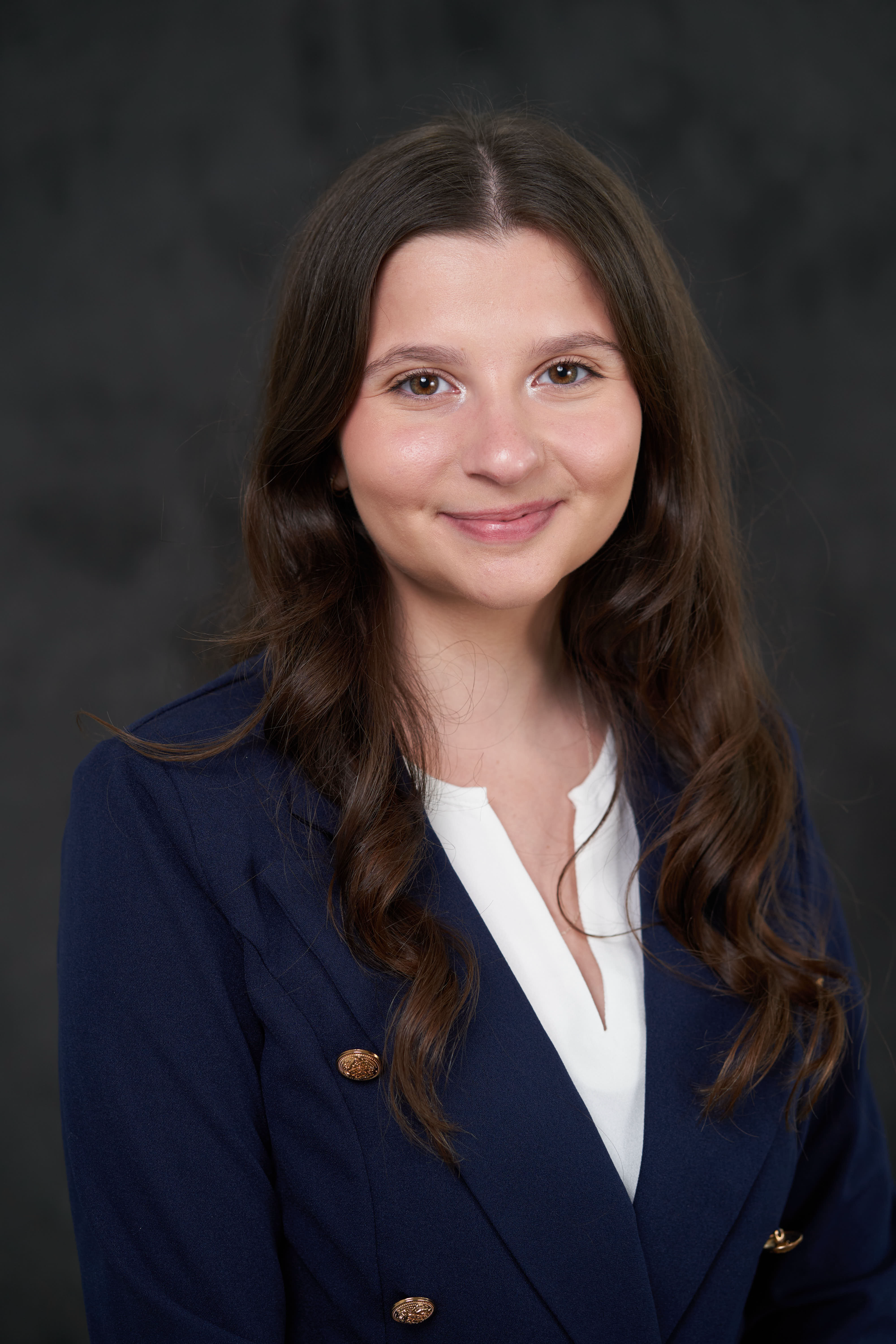 Professional headshot of a young woman wearing a navy blazer with gold buttons and white blouse against a dark background