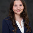 Professional headshot of a young woman wearing a navy blazer with gold buttons and white blouse against a dark background