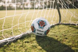 Wilson NCAA soccer ball resting on grass inside a soccer goal net on a sunny field
