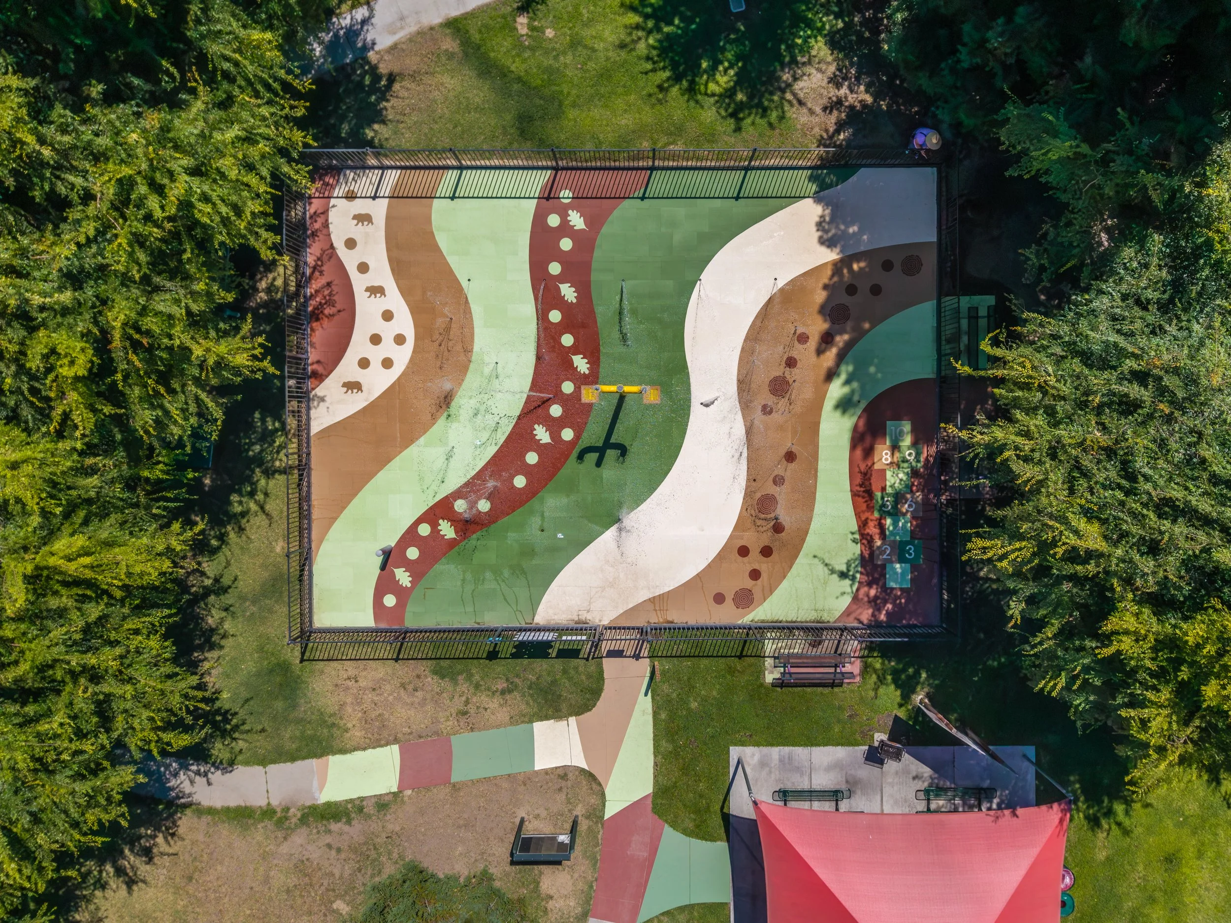 Aerial view of a colorful playground with curved pathways, sand areas, and green spaces surrounded by trees