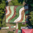 Aerial view of a colorful playground with curved pathways, sand areas, and green spaces surrounded by trees
