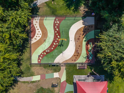 Aerial view of a colorful playground with curved pathways, sand areas, and green spaces surrounded by trees