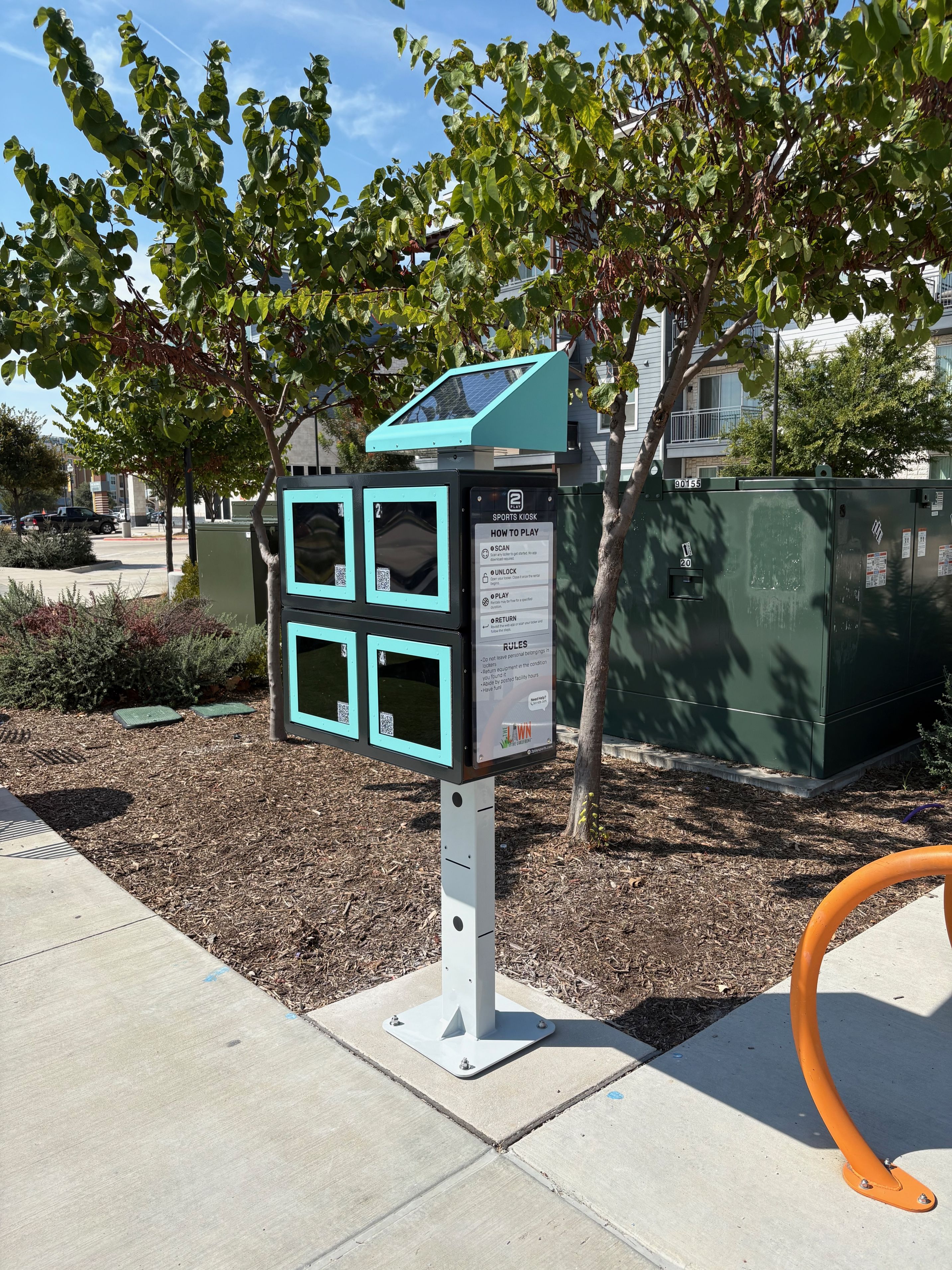 Turquoise and black interactive kiosk with four window panels in a shaded urban park area surrounded by trees and landscaping.