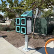 Turquoise and black interactive kiosk with four window panels in a shaded urban park area surrounded by trees and landscaping.