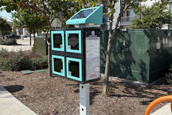 Turquoise and black interactive kiosk with four window panels in a shaded urban park area surrounded by trees and landscaping.