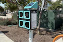 Turquoise and black interactive kiosk with four window panels in a shaded urban park area surrounded by trees and landscaping.