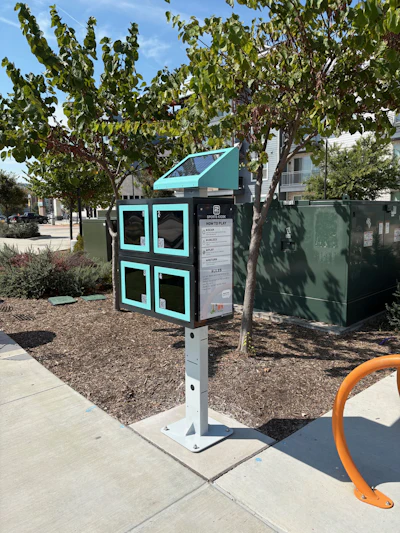 Turquoise and black interactive kiosk with four window panels in a shaded urban park area surrounded by trees and landscaping.