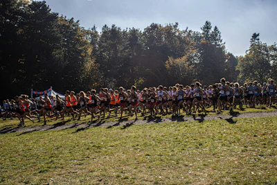 Large group of runners gathered on a grassy field during an outdoor race event with forest trees in background