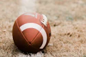 Brown football with white stripes resting on grass and dirt ground