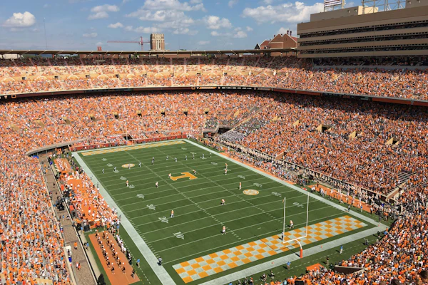 Packed football stadium with orange-clad fans, green field, and yellow end zone during game day