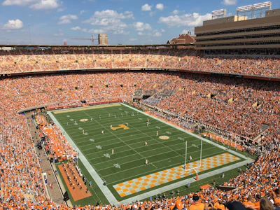 Packed football stadium with orange-clad fans, green field, and yellow end zone during game day
