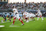 High school football game in progress with players in light blue and red uniforms competing on field during daytime match.