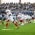 High school football game in progress with players in light blue and red uniforms competing on field during daytime match.