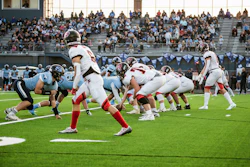 High school football game in progress with players in light blue and red uniforms competing on field during daytime match.