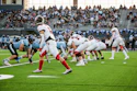High school football game in progress with players in light blue and red uniforms competing on field during daytime match.